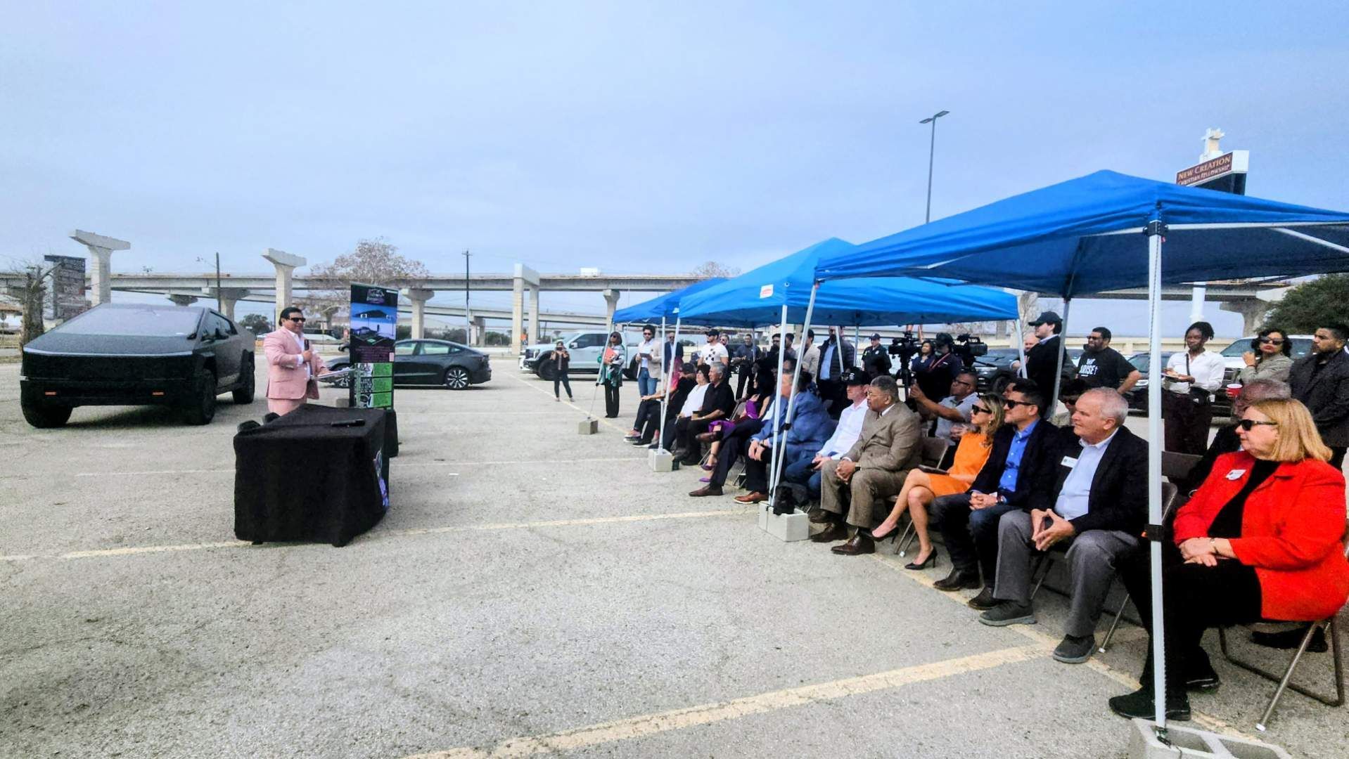 People seated under blue canopies at an outdoor event, with a speaker at a podium nearby.
