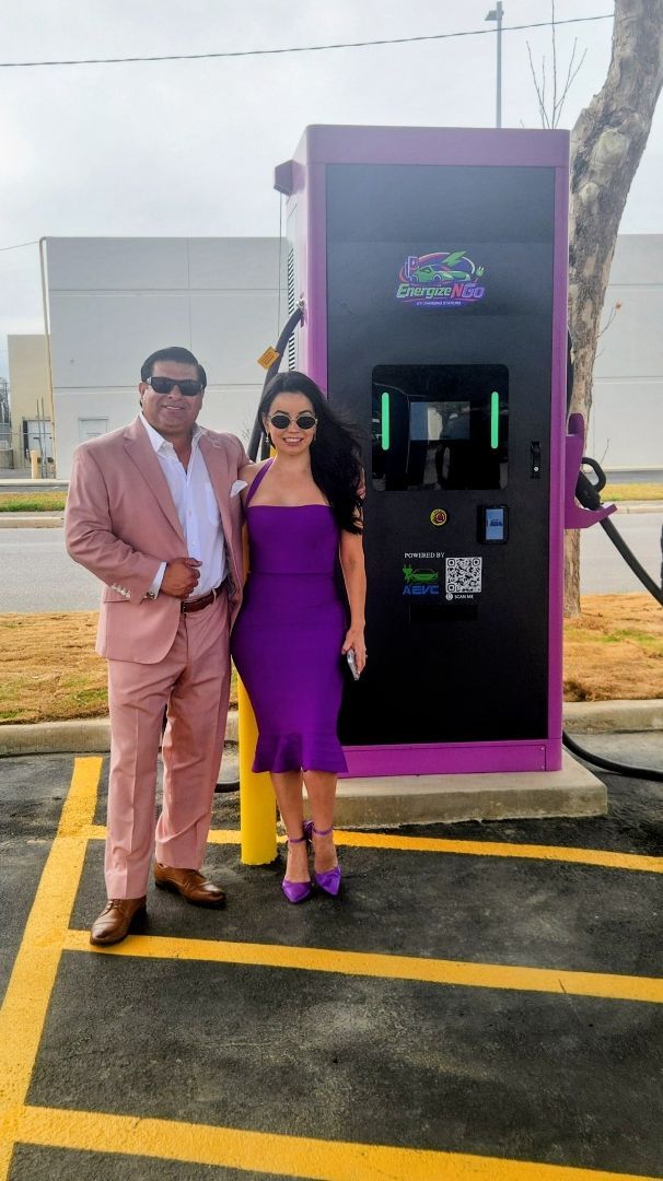 Two people posing beside a purple charging station in a parking lot, one in a pink suit and one in a purple dress.