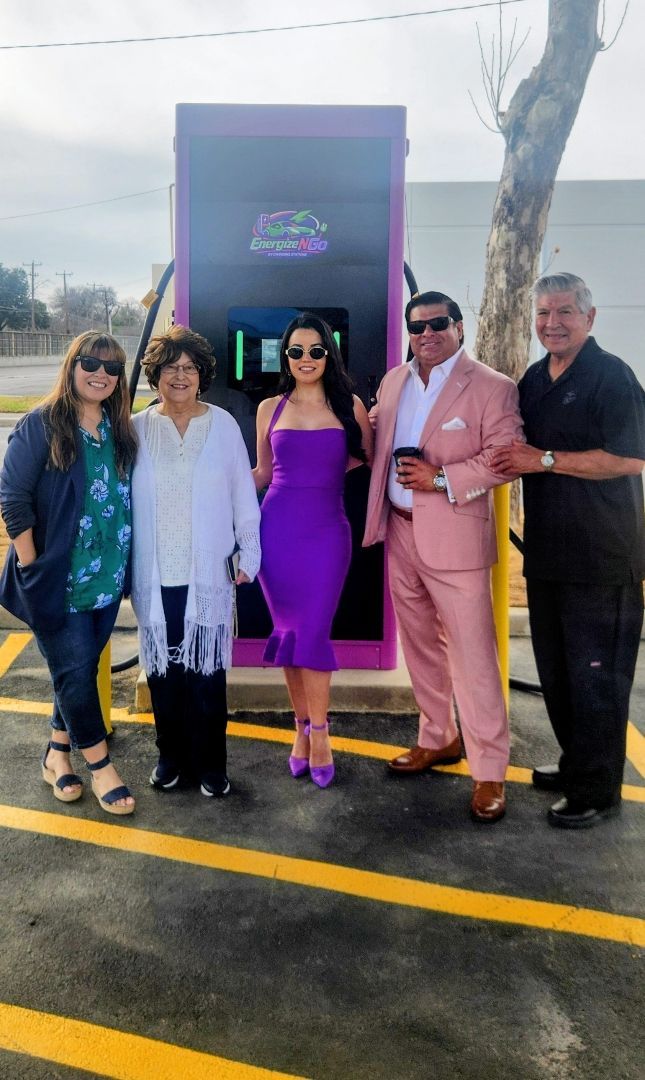 Five people posing outdoors on a wet pavement in front of a purple-lit sign and palm trees.