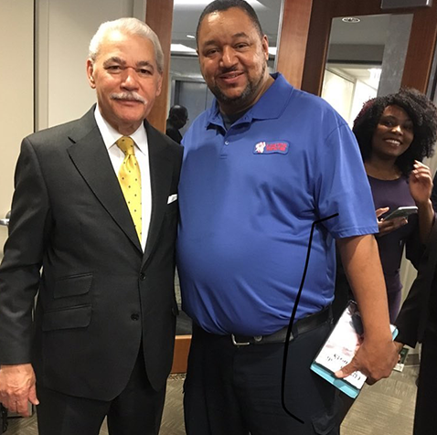 A man in a suit and tie poses with another man in a blue shirt