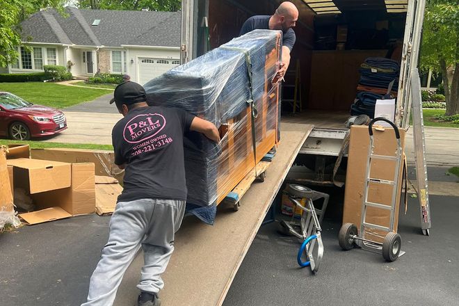 Two movers loading a wrapped pallet onto a truck ramp in a residential driveway