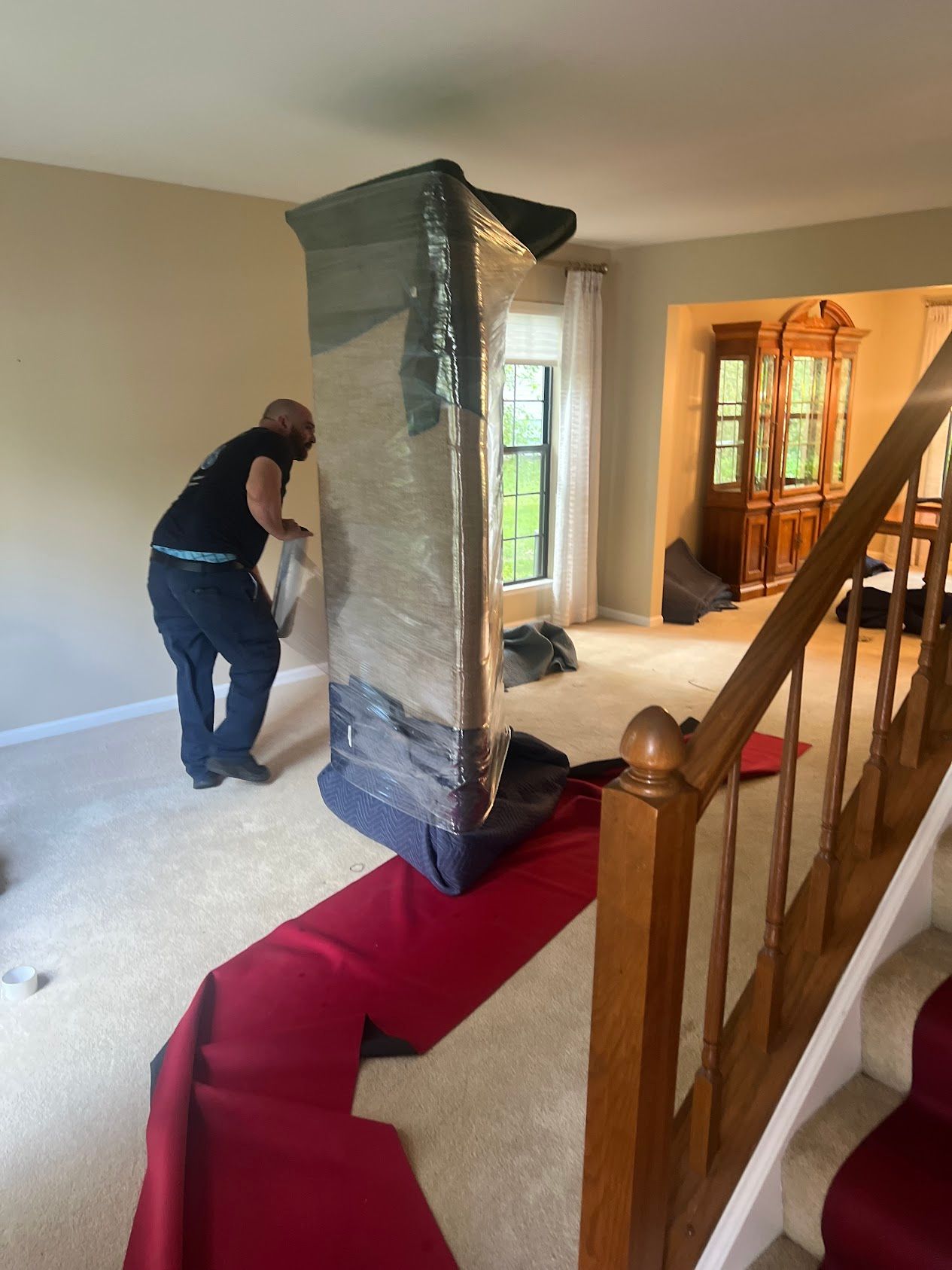 Mover wrapping a large mirror in a beige living room near a staircase with red carpet runner