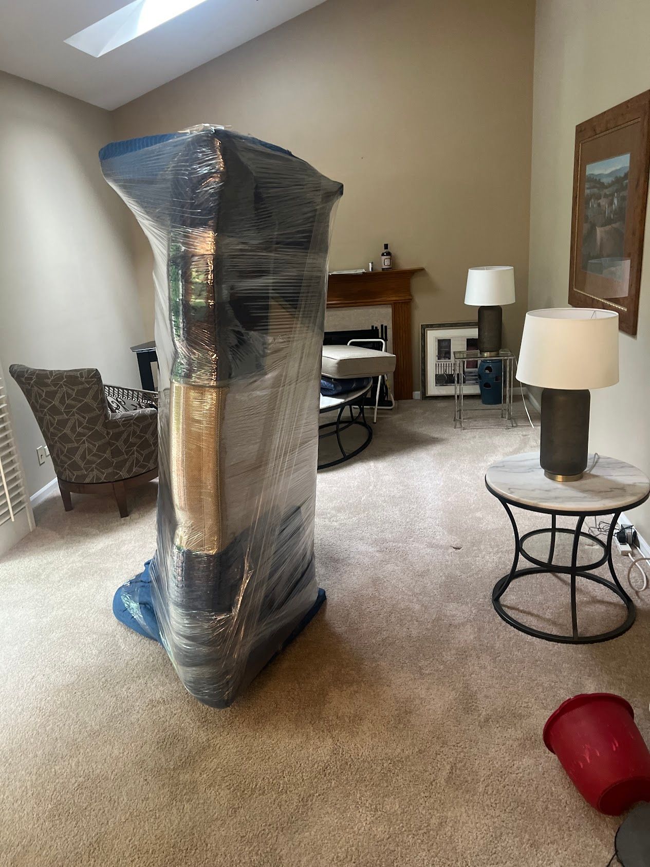 Wrapped tall furniture piece in a carpeted room with tables, lamps, and a chair