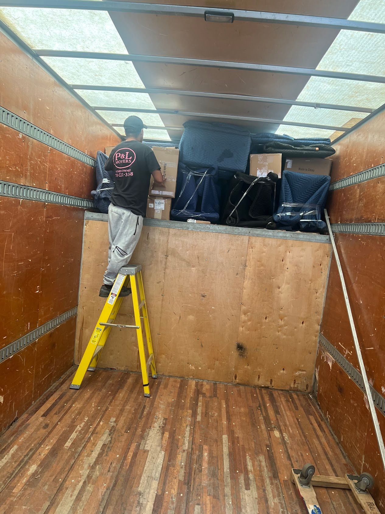 Worker on a yellow ladder securing black items inside an empty wood-paneled cargo trailer.