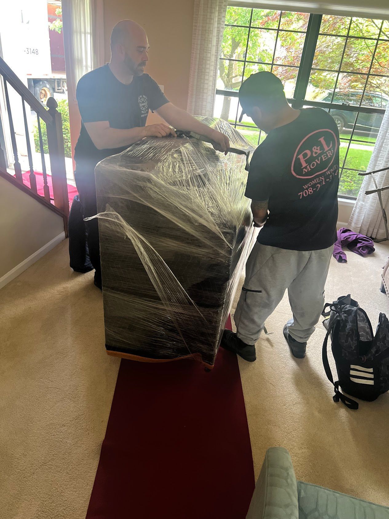 Two people moving a wrapped sofa through a doorway onto a red carpet in a bright entryway.