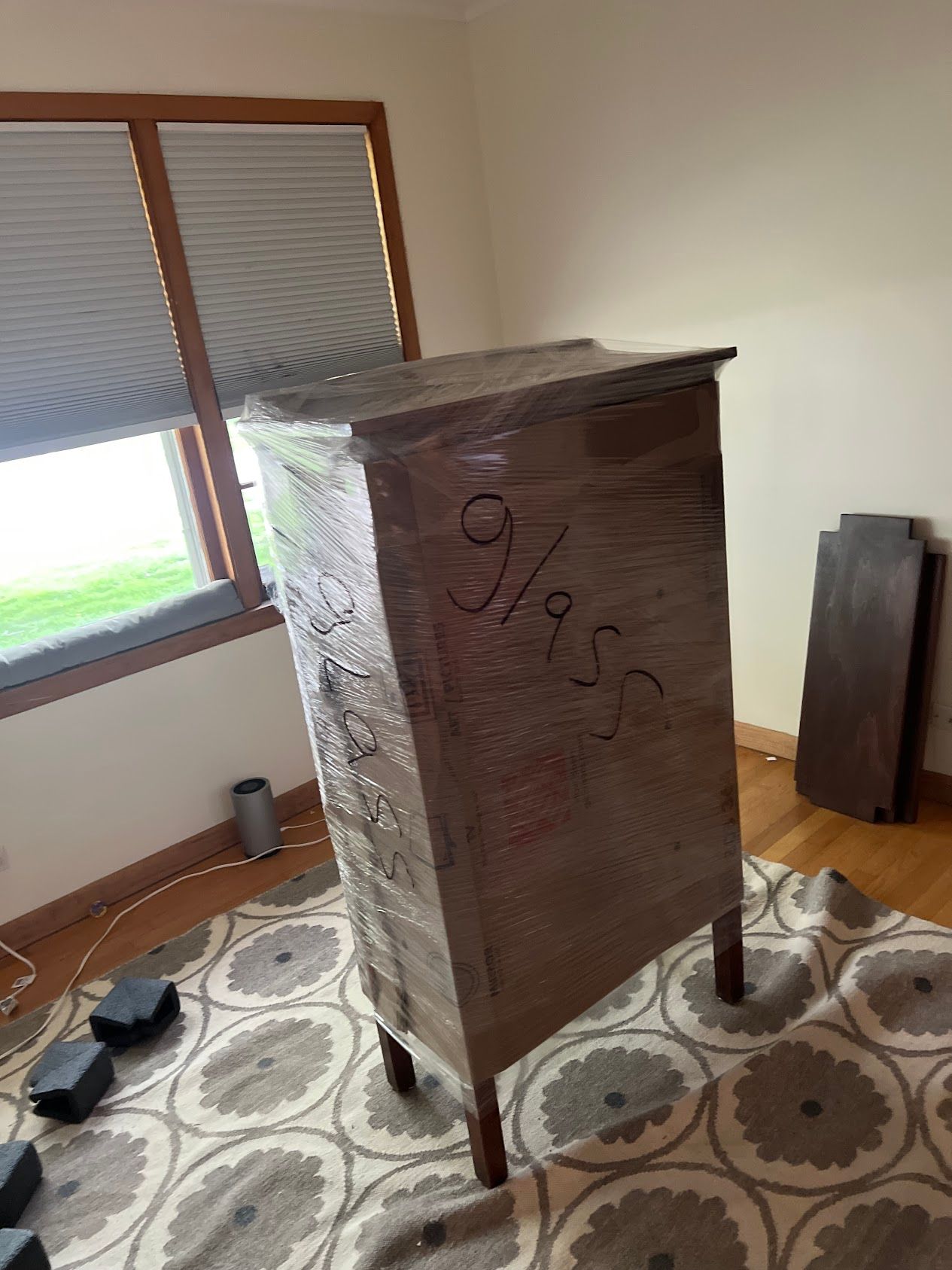Large wrapped wooden cabinet with scribbled markings in a room by a window, on a patterned rug.