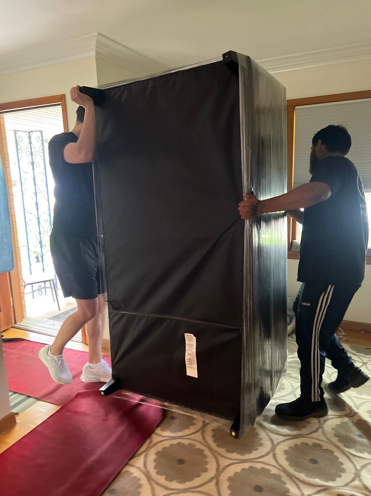 Two people carrying a large black mattress through a doorway in a living room