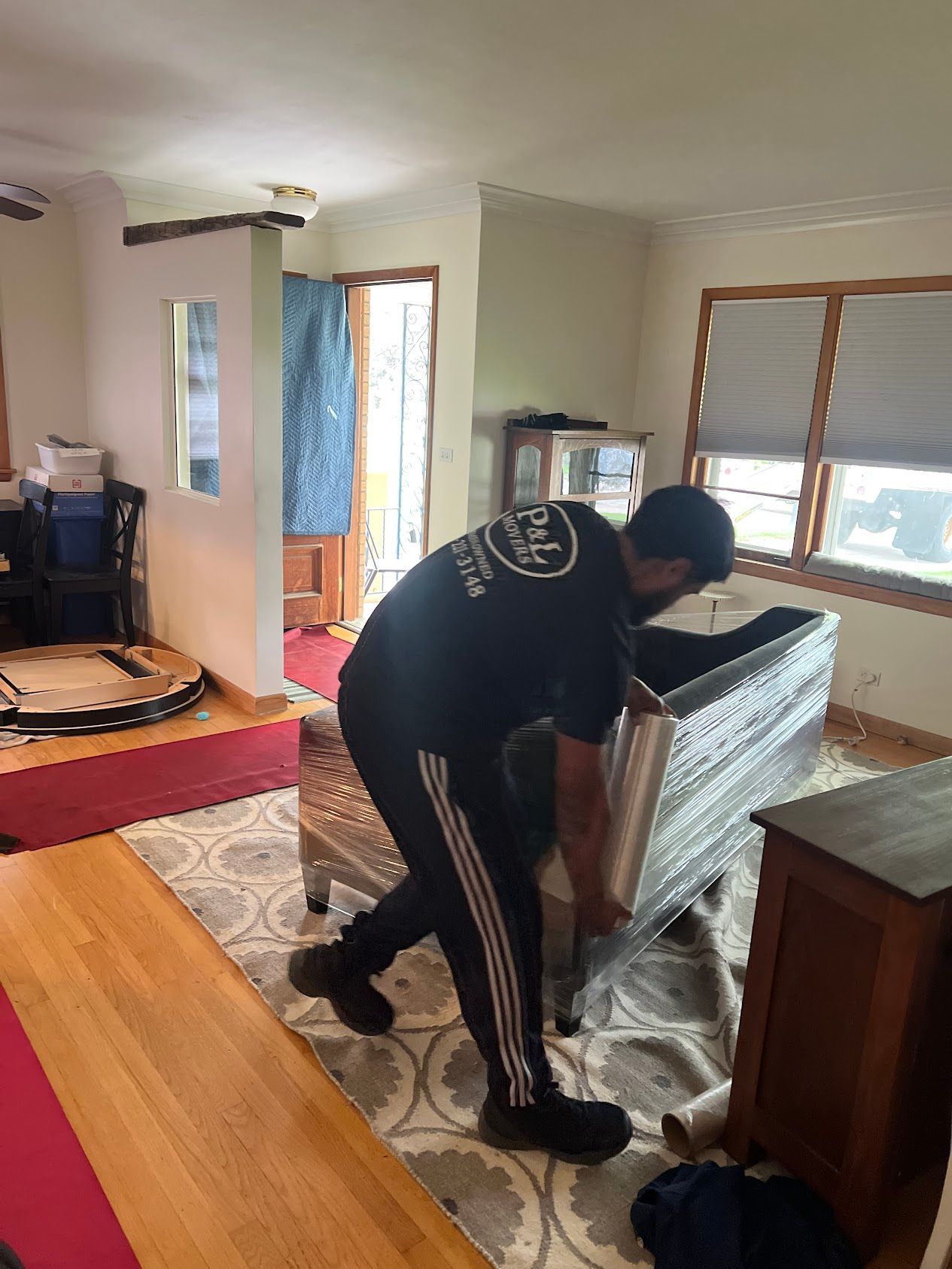 Person moving a wrapped mattress in a living room with wooden floors and large windows