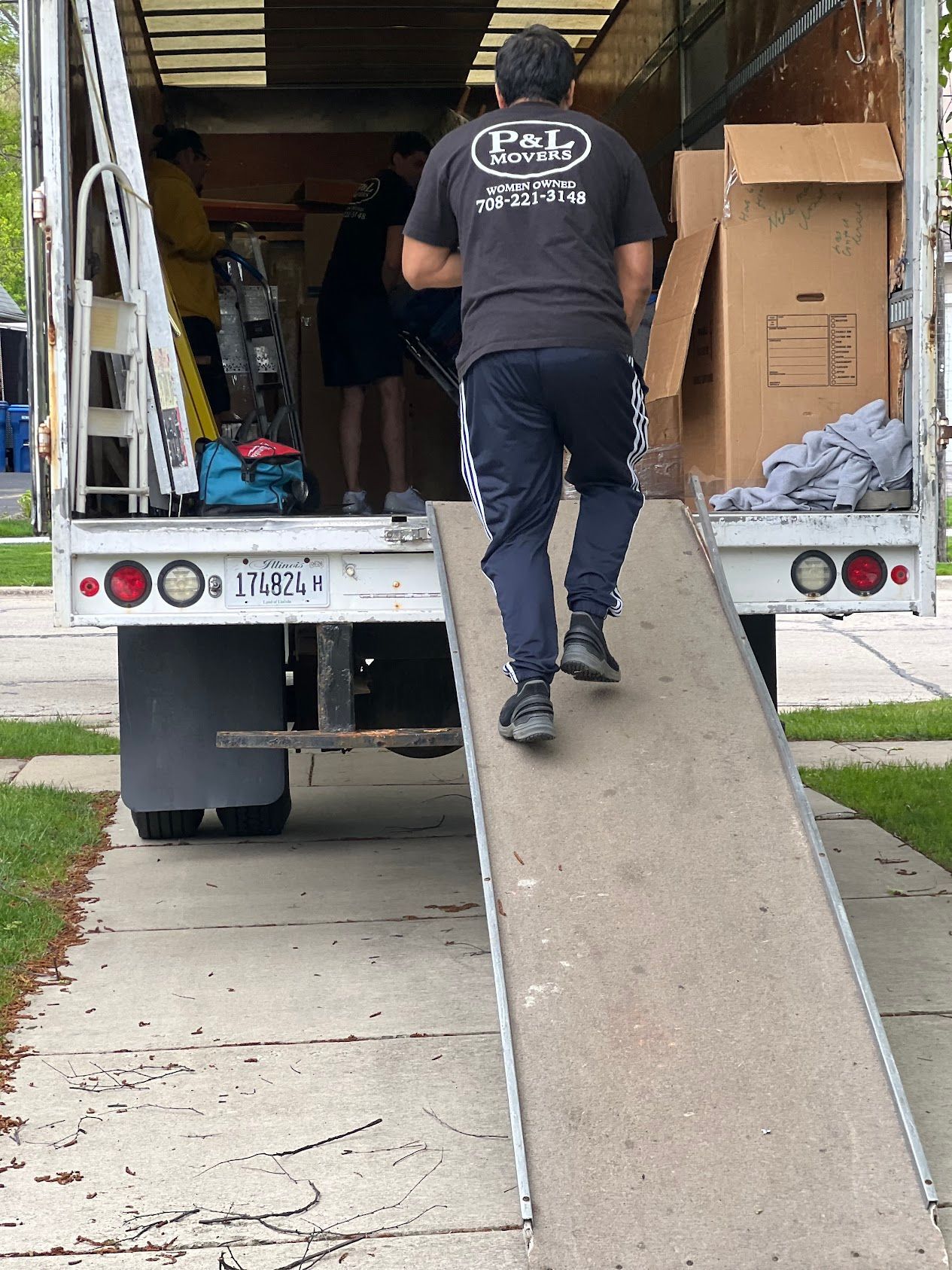 Worker pushing a hand truck up a ramp into a moving truck parked by a sidewalk