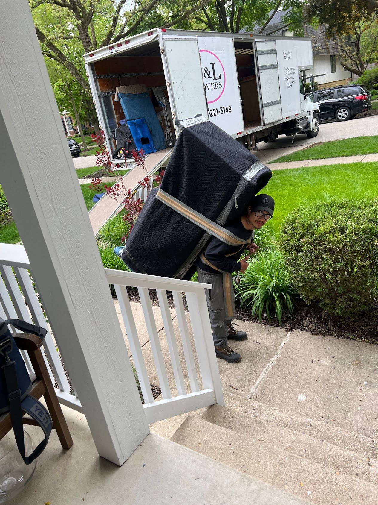 Person carrying a large black mattress up a front porch steps near a grassy yard
