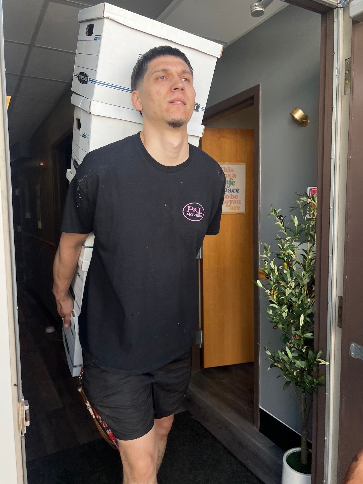 Man standing in a doorway, wearing a black T-shirt and shorts, with boxes stacked behind him.