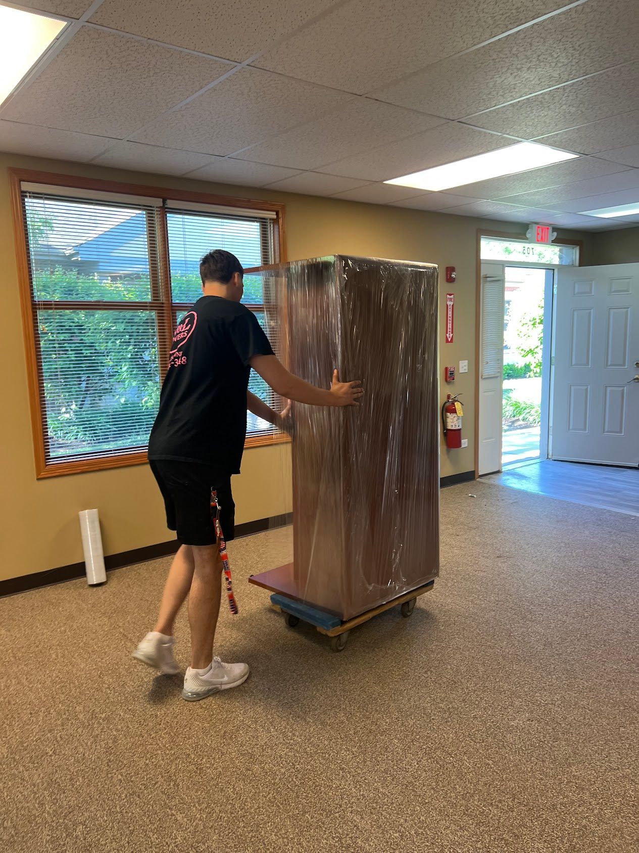 Person pushing a large wooden cabinet on a dolly in a carpeted room with windows and an open door.