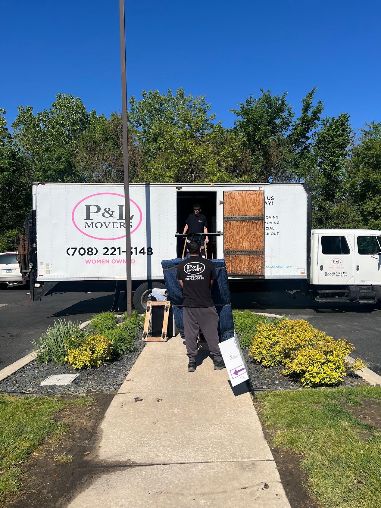 Person carrying a folded sign toward a white box truck with boarded windows on a sunny sidewalk