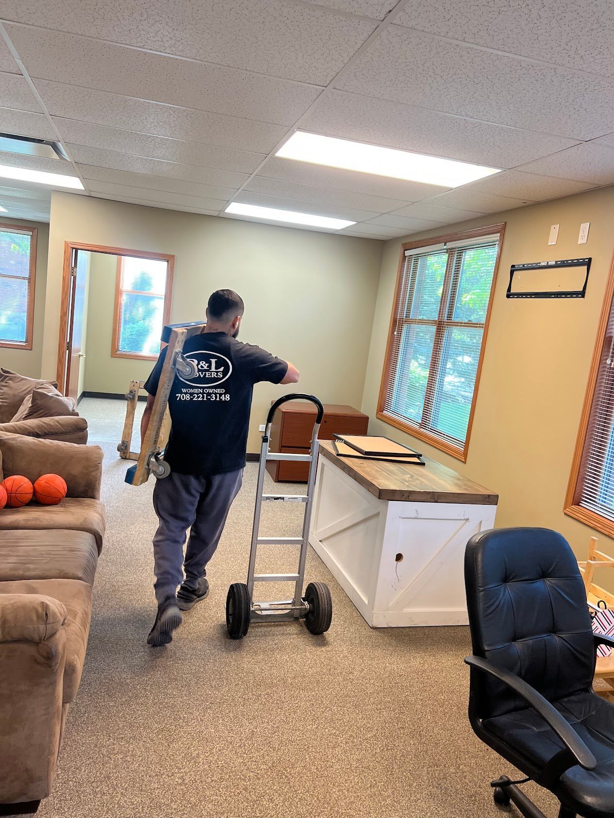 Person pushing a hand truck through a beige living room with furniture and windows