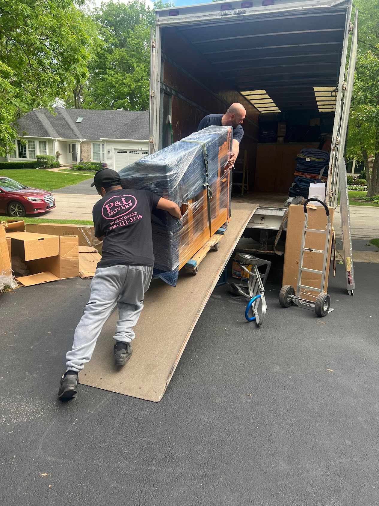 Workers loading a large wrapped item down a ramp into a moving truck in a driveway
