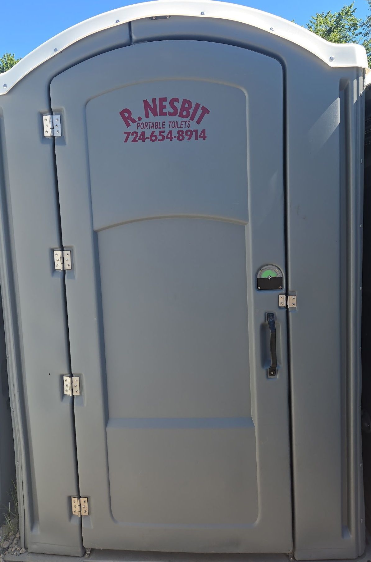 Gray portable toilet with arched top, door closed. R. NESBIT phone number on door.