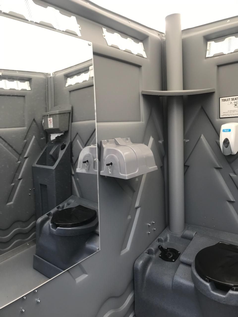Interior of a gray portable toilet, showing a toilet, sink, mirror, and soap dispenser.