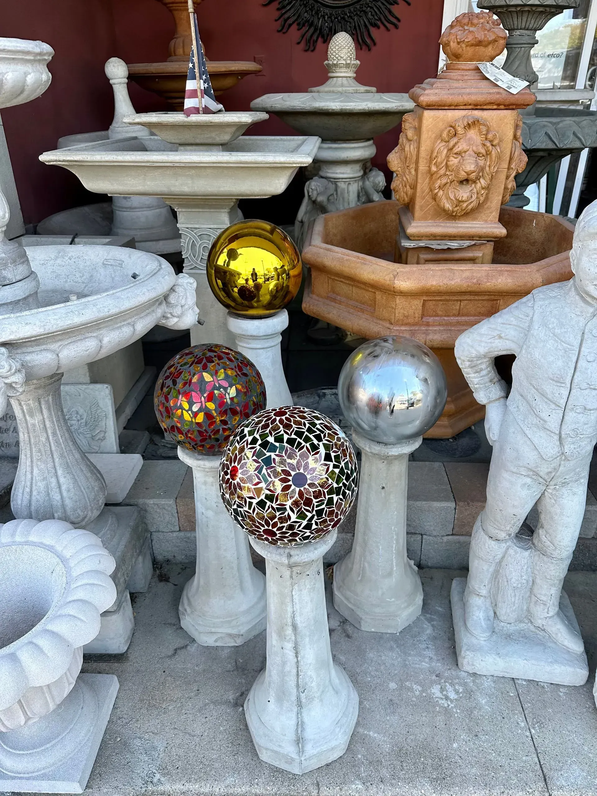 A display of various garden ornaments, including concrete birdbaths, pedestals with decorative spheres, and a statue.
