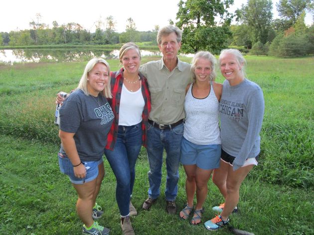 A group of five people stand smiling together in a grassy field in front of a pond at sunset.
