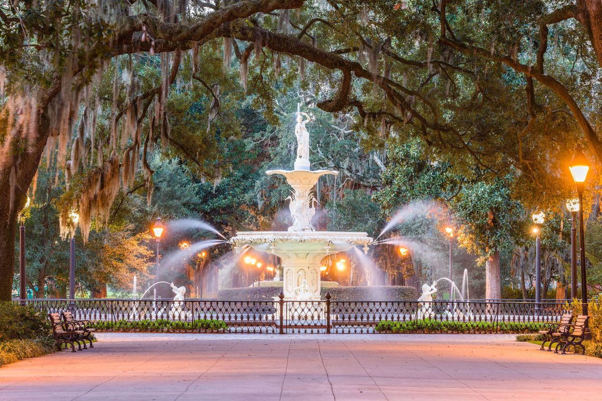 The ornate white Forsyth Park fountain in Savannah, Georgia, glows at dusk surrounded by moss-draped trees.