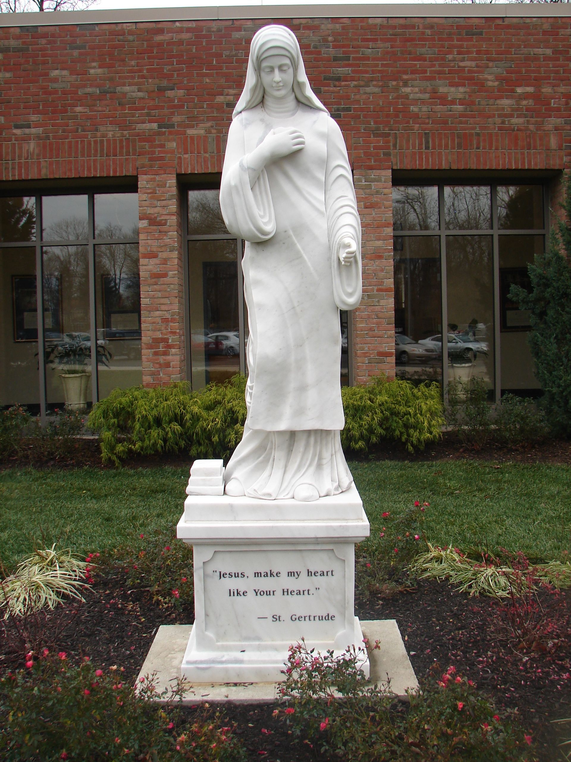 A white stone statue of a woman in a veil stands on a pedestal inscribed with text in front of a brick building.