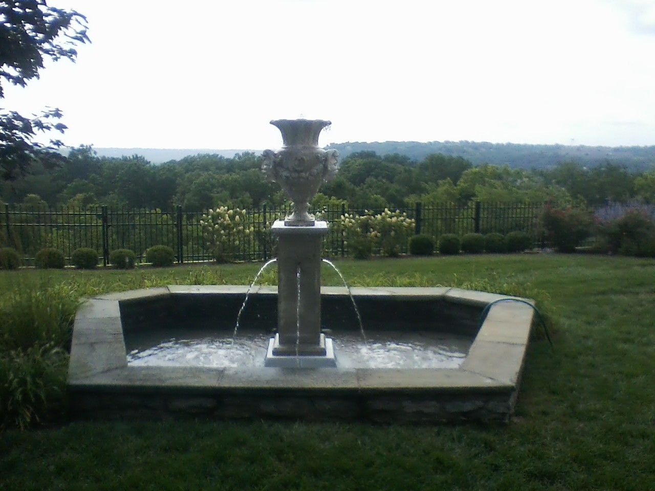 A stone water fountain with an urn-shaped top stands in a grassy backyard overlooking a distant wooded landscape.