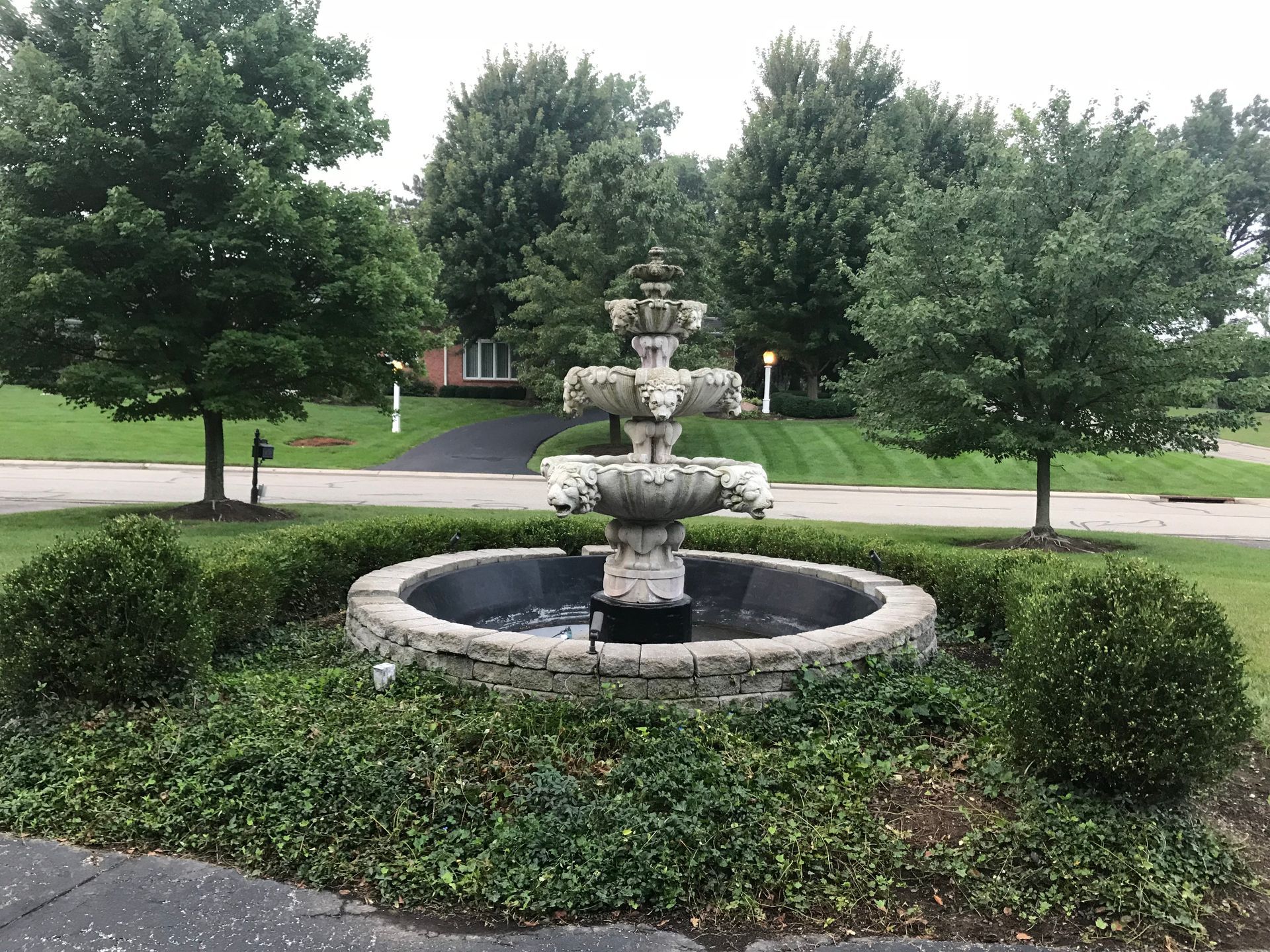 A three-tiered stone fountain stands in the center of a circular landscaped garden surrounded by green trees and grass.