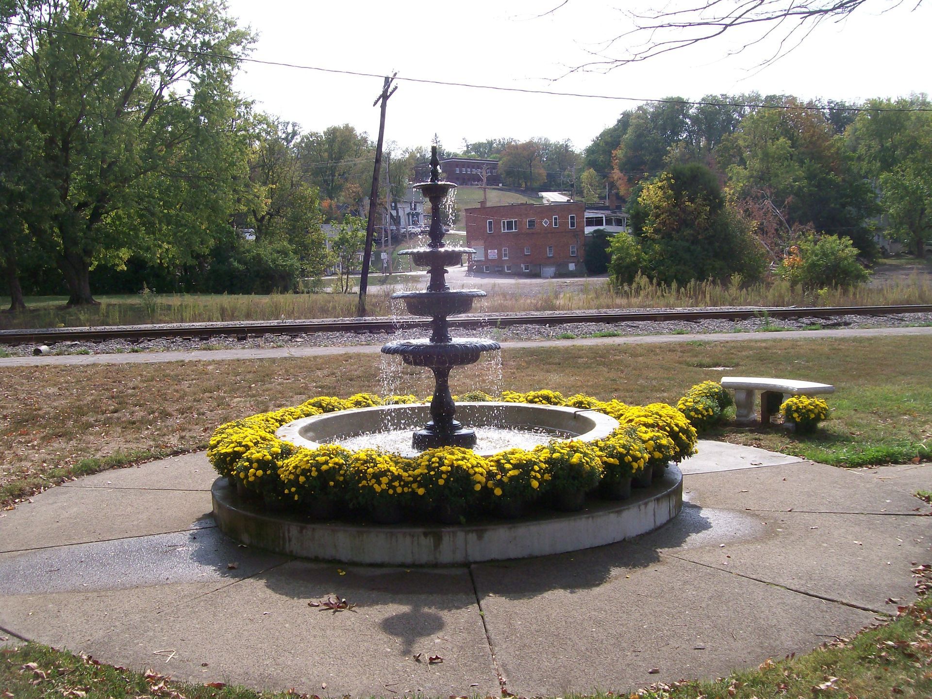 A three-tiered black fountain surrounded by yellow flowers in a park, with railroad tracks in the background.