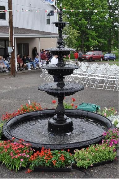 A tiered, black metal fountain sits outdoors, surrounded by colorful potted flowers, with chairs and people in the back.