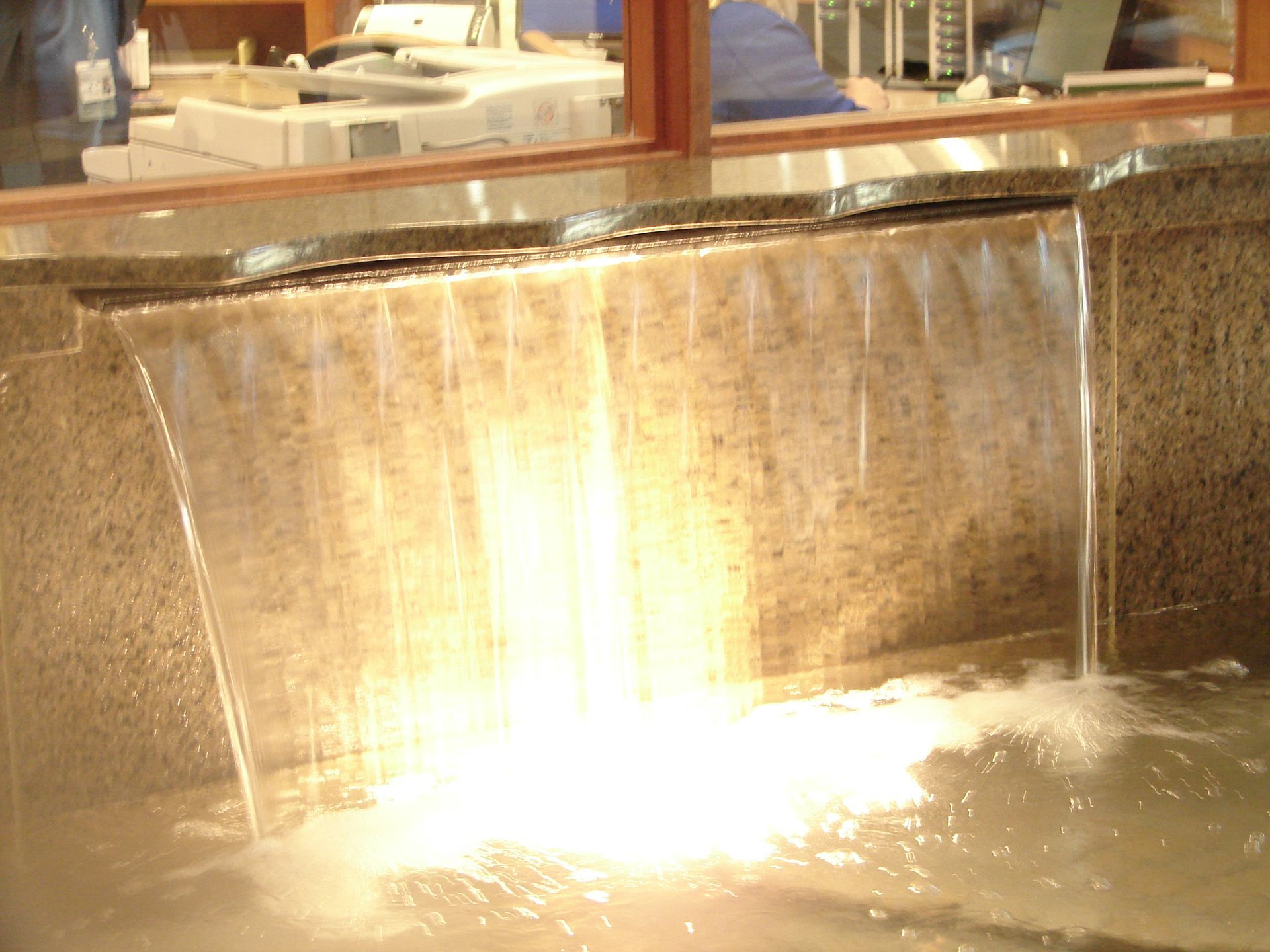 A sheet of water flows down a polished granite stone wall into a basin, creating a splash at the base.