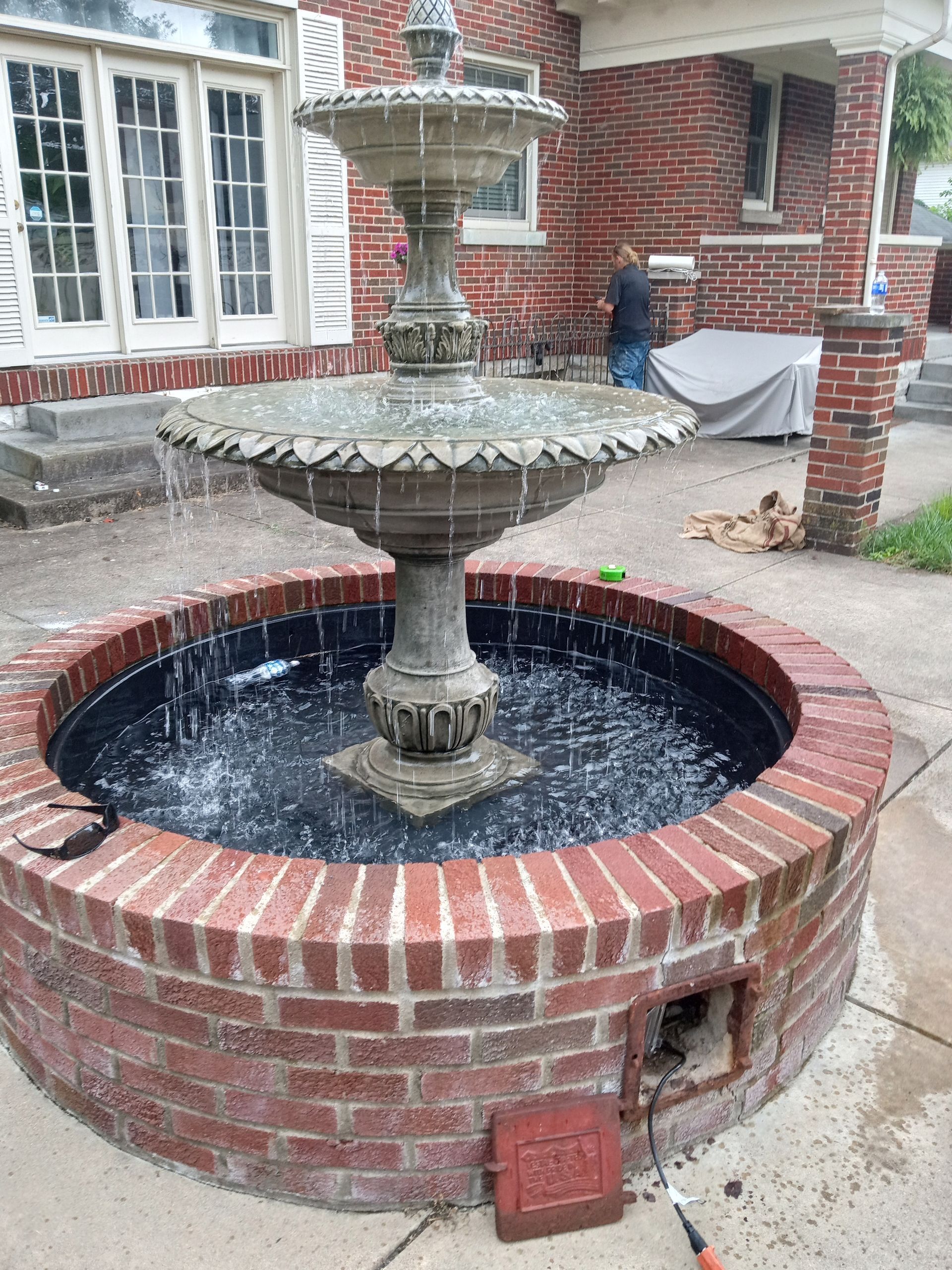 A tiered stone fountain flows inside a circular brick basin on a patio in front of a red brick house.
