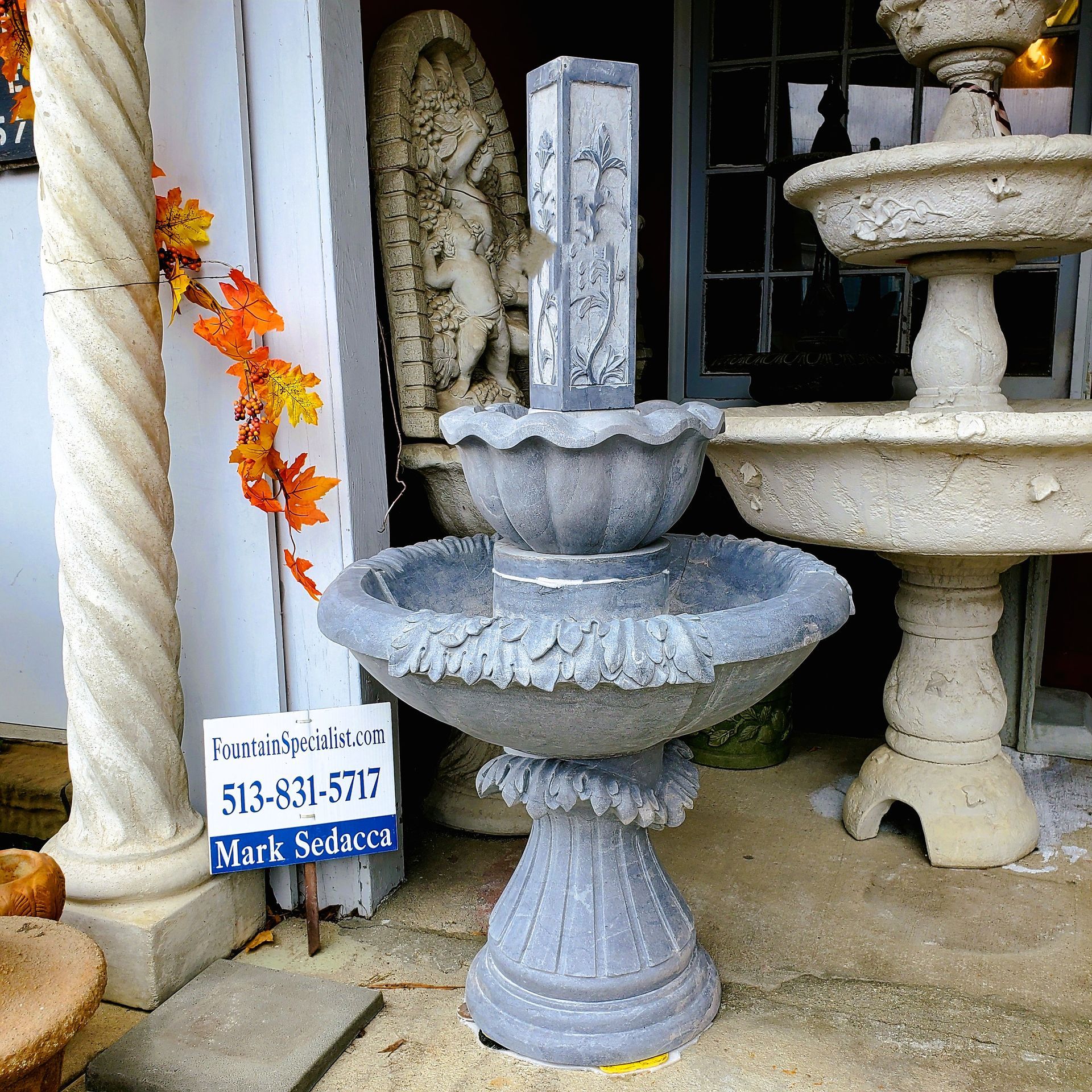 A multi-tiered gray stone fountain stands outdoors next to a spiral column and a sign with a phone number.