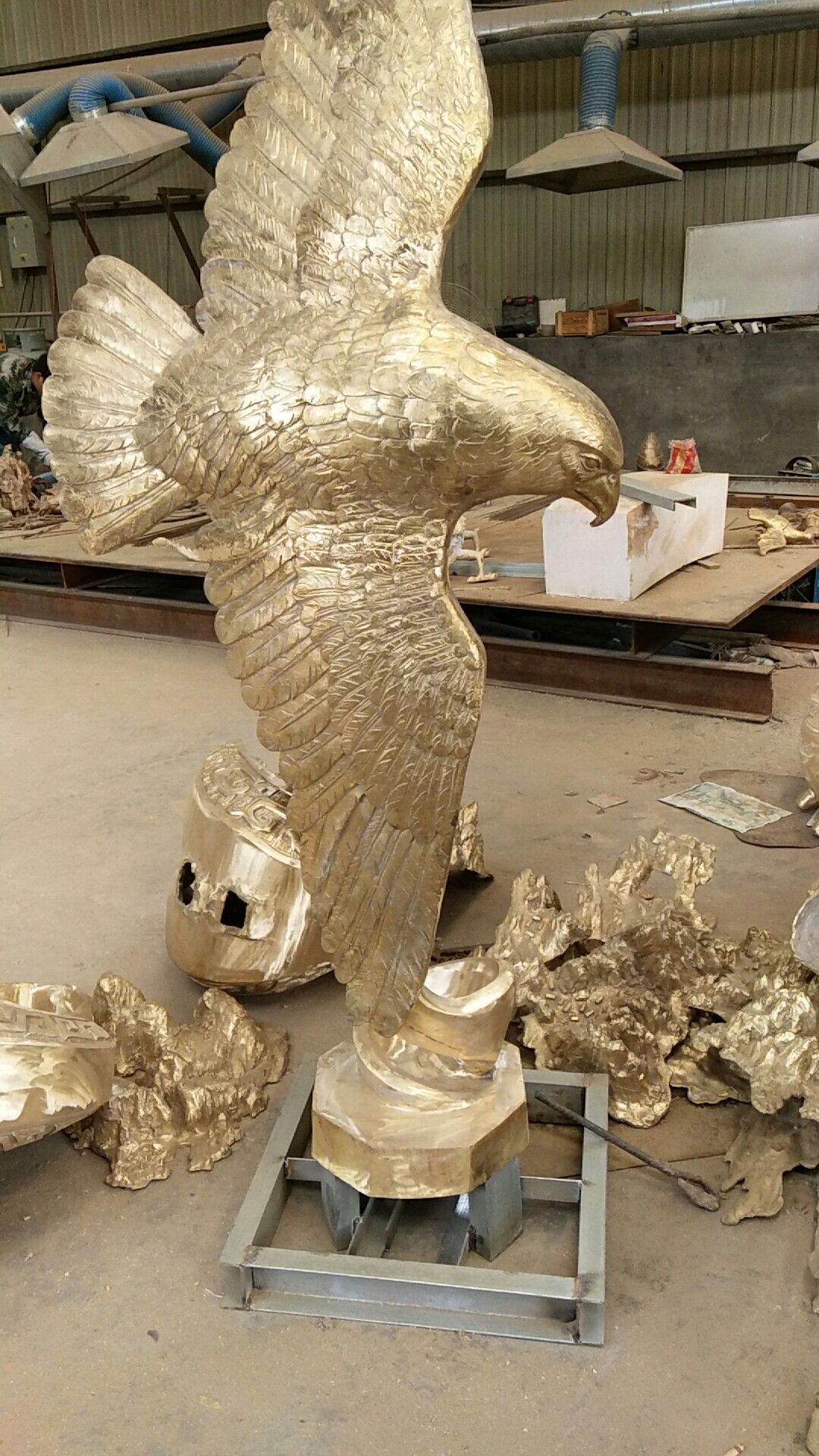 A golden eagle statue stands on a metal base in a workshop, surrounded by scattered, unpolished golden metal fragments.