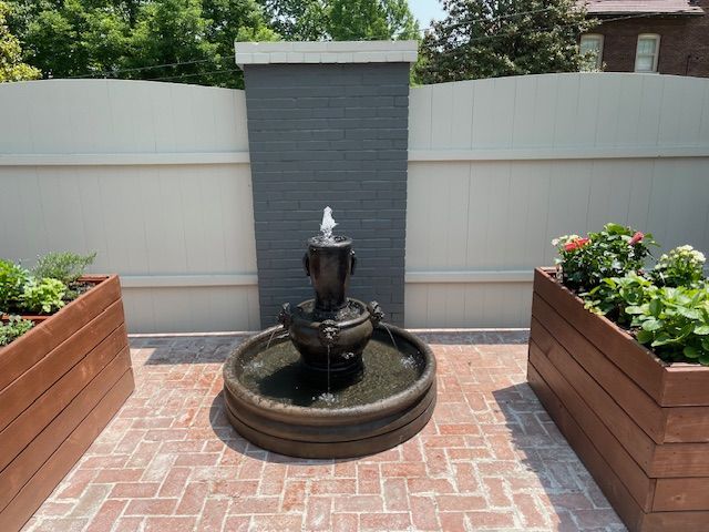 A tiered outdoor fountain stands on a brick patio between two wooden planters, all set against a white and gray fence.