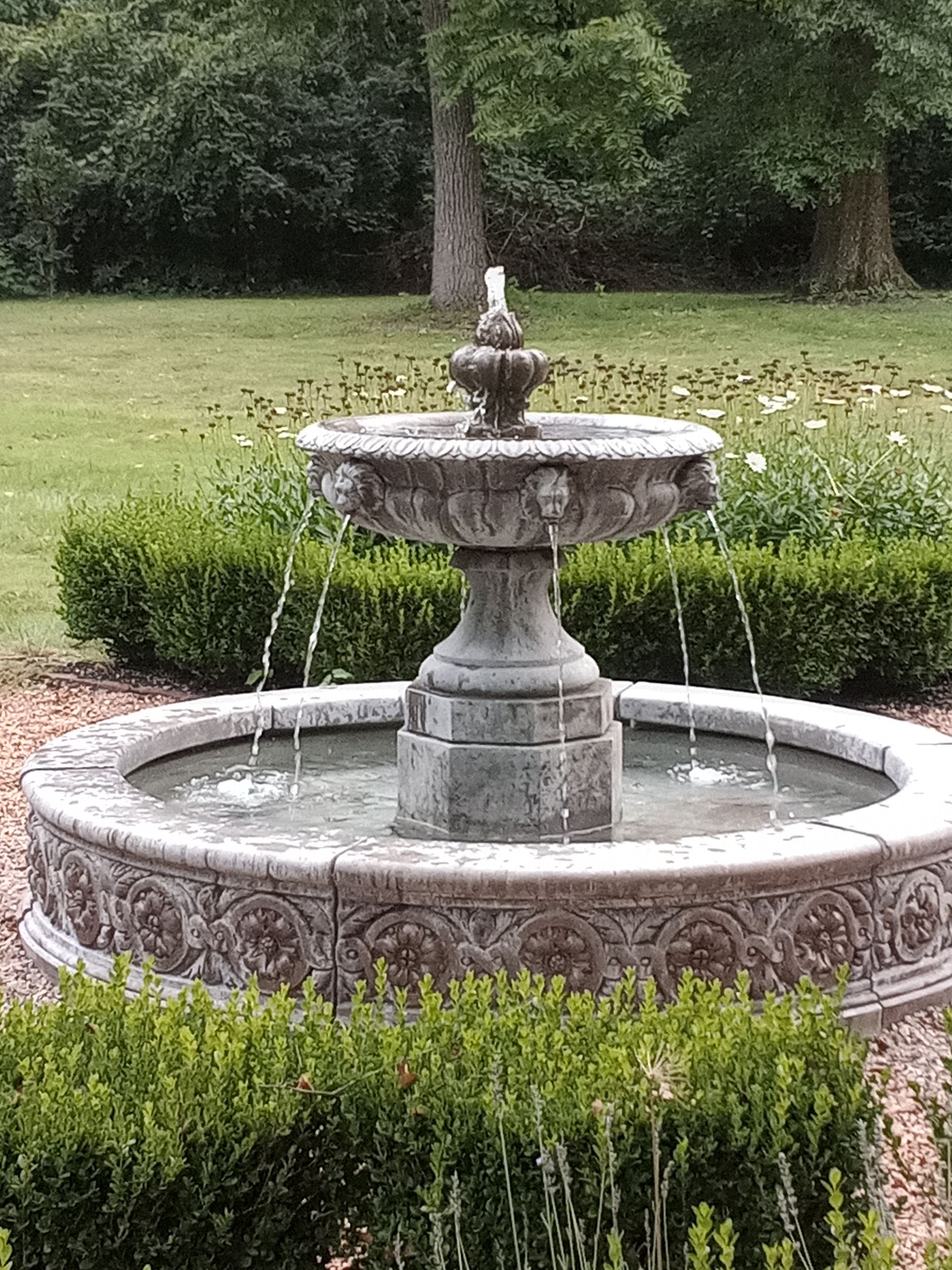 A decorative stone fountain with cascading water, set in a garden with neatly trimmed hedges and trees in the background.