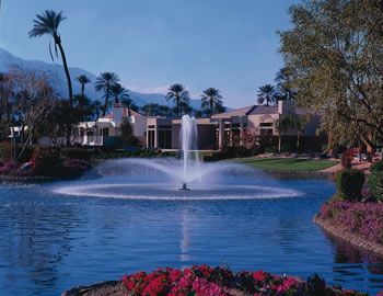 A fountain sprays water in the center of a calm lake, surrounded by palm trees, lush landscaping, and a distant building.