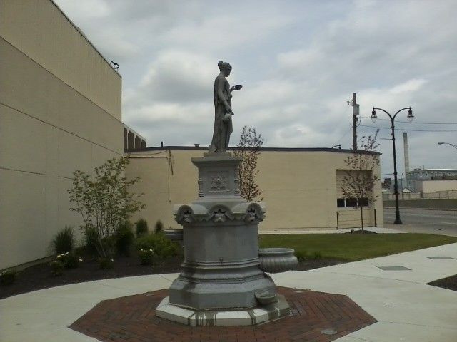 A metal fountain featuring a standing figure holding an object, situated on a brick plaza next to a beige building.