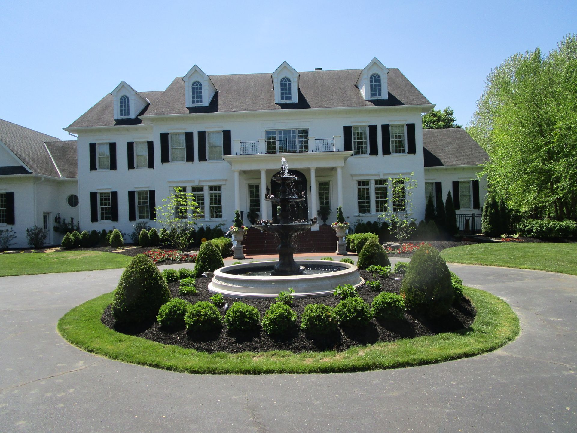A large white colonial-style home with black shutters, a circular driveway, and a tiered stone fountain in the center.