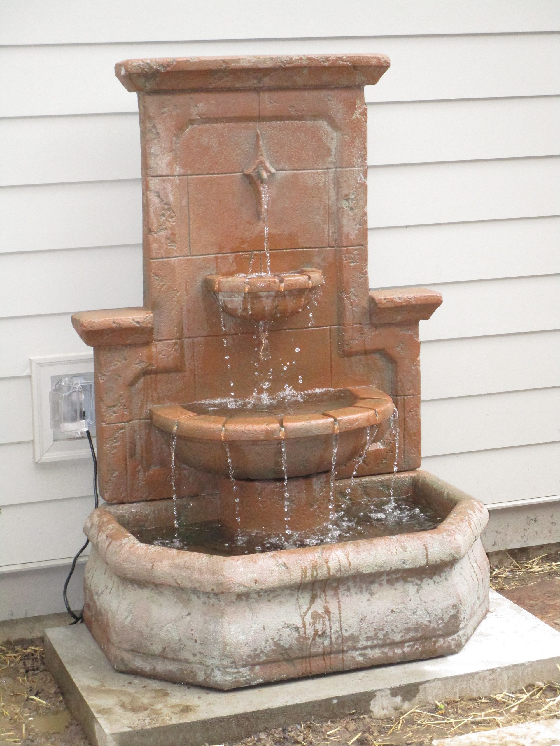 A tiered terracotta-colored wall fountain with cascading water, situated on a concrete base against a white wall.