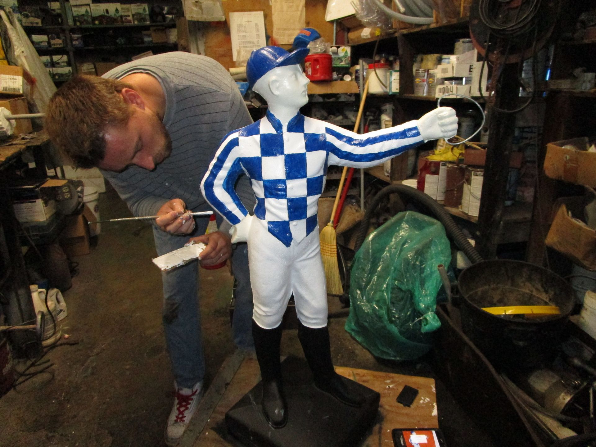 A person paints a blue-and-white checkered jockey statue in a cluttered workshop.