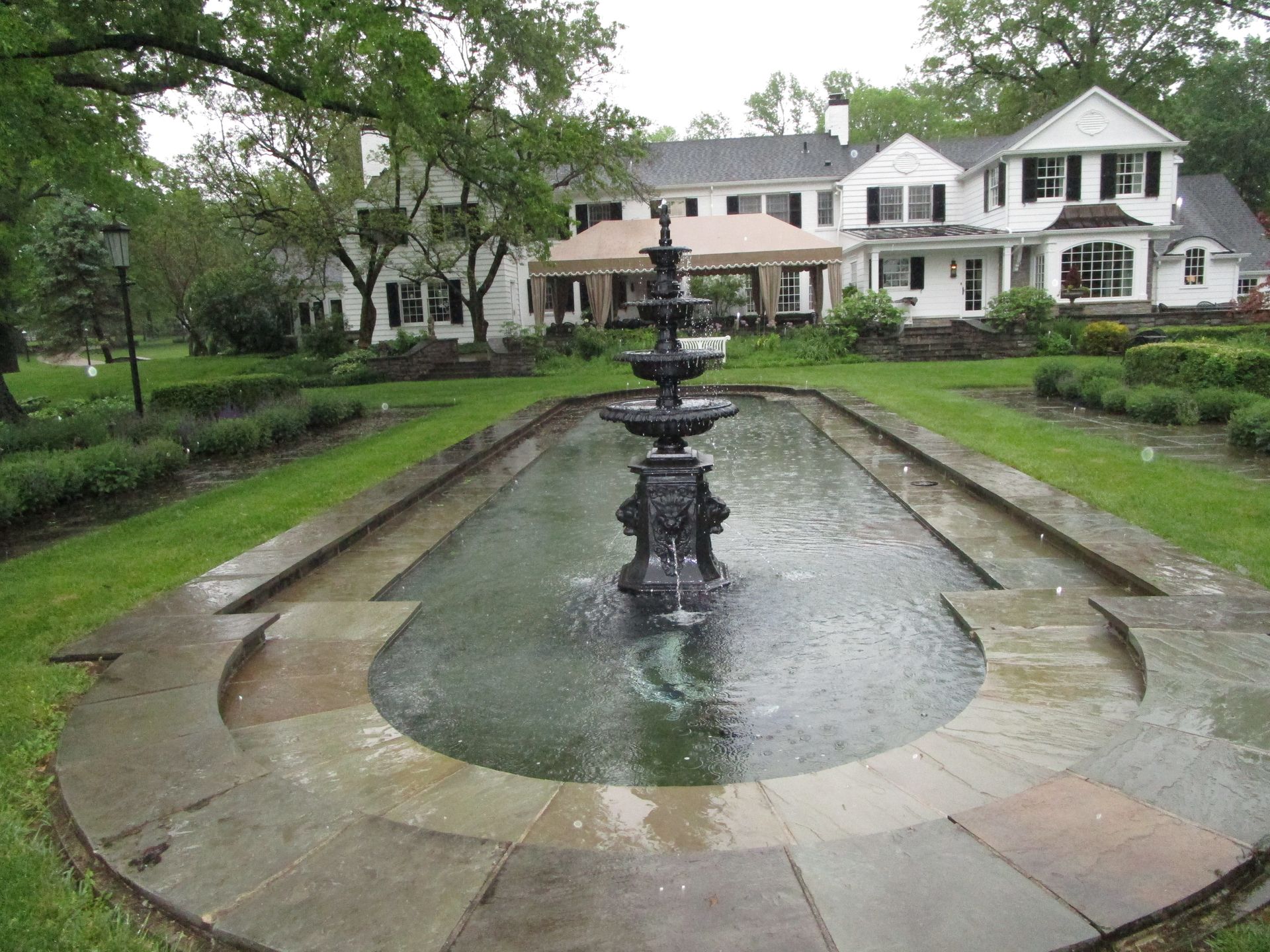 A tiered stone fountain sits in a rectangular pool surrounded by green lawn in front of a white house on a rainy day.