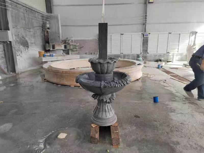 A tiered stone fountain assembly sits on wooden blocks in a workshop, with a large stone basin in the background.