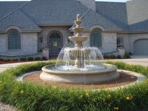 A three-tiered stone fountain sits in the center of a circular garden bed in front of a gray stone house.