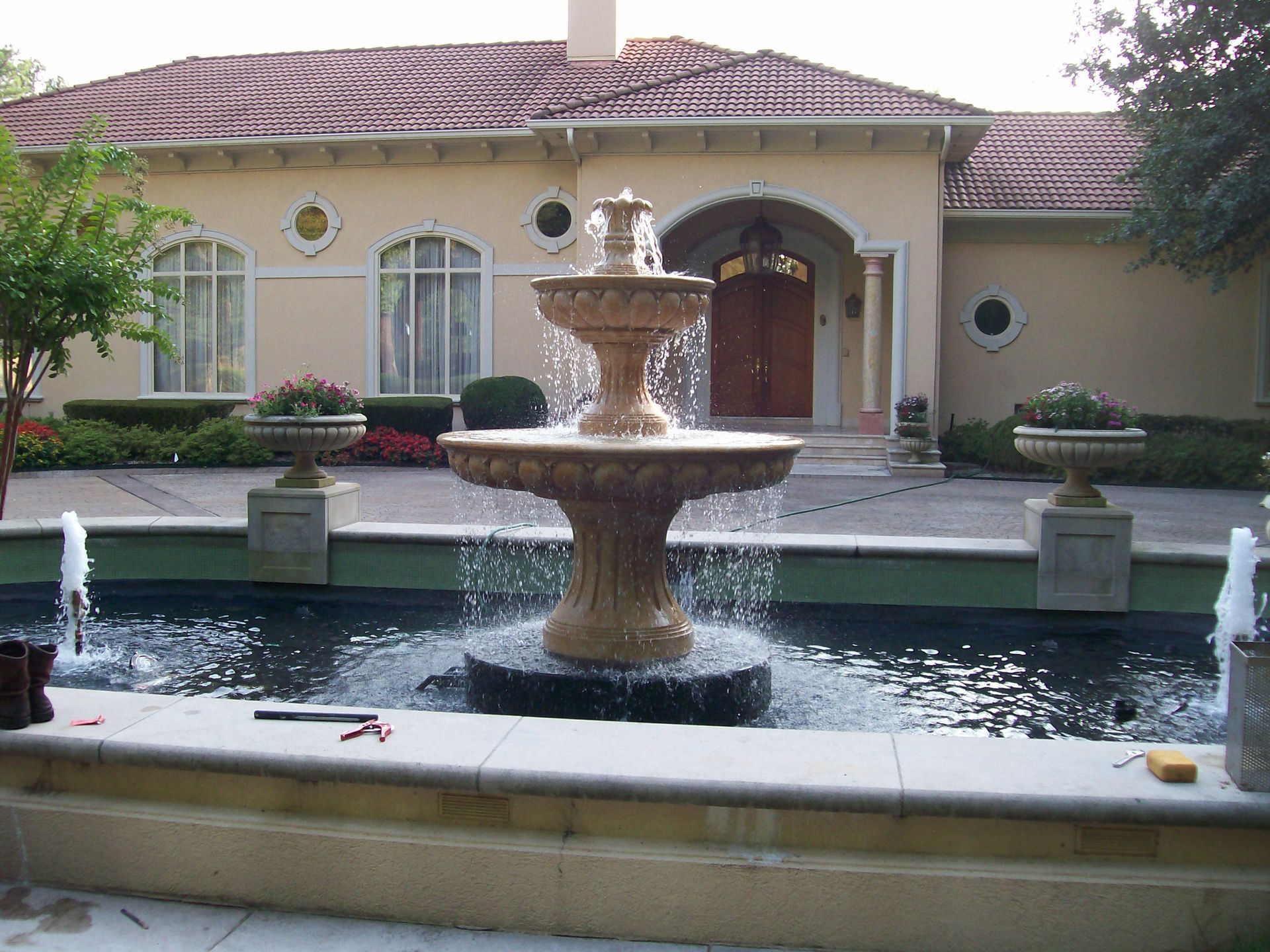 A multi-tiered stone fountain splashes in front of a tan, single-story house with arched windows and a red-tiled roof.