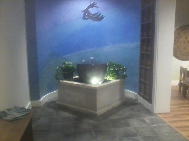 A lit, bowl-shaped fountain centered in a stone base against a blue, mural-painted wall in a room with tiled flooring.