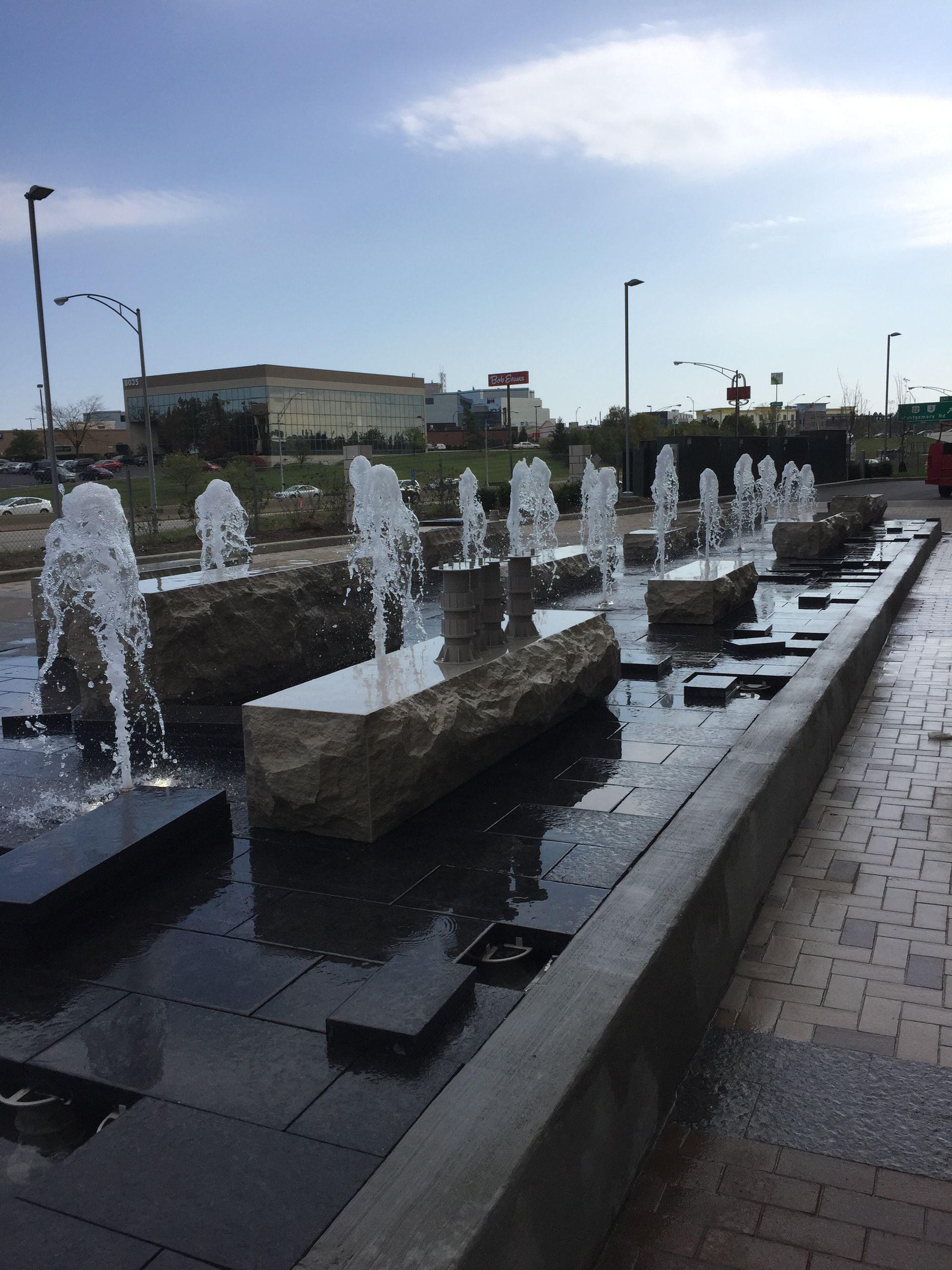 A row of stone blocks in a long fountain, with water spraying upward into the air under a bright blue sky.
