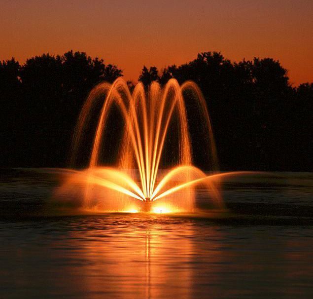 A fountain illuminated in golden light shoots water into an arc against a sunset sky over a dark lake.