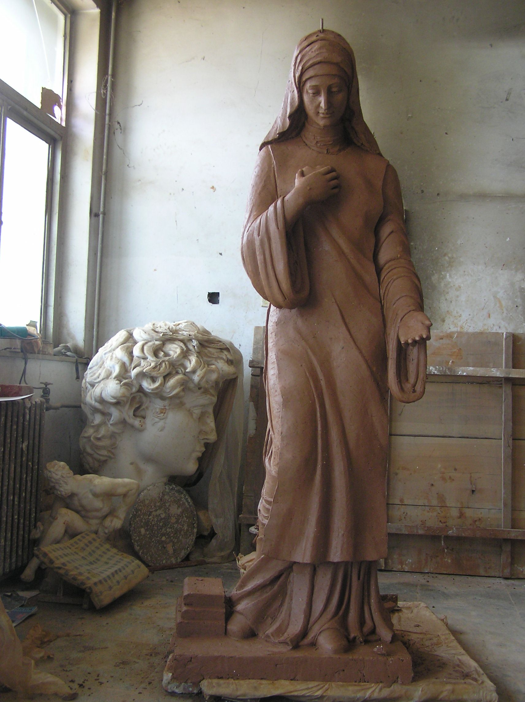 A clay statue of a robed figure standing in a studio, with a large plaster bust visible in the background.