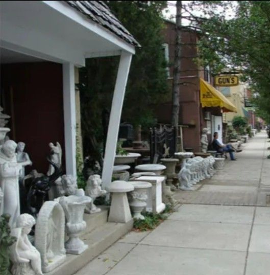 Outdoor shop display featuring various concrete statues and urns on a sidewalk, with a person resting near a yellow awning.