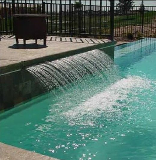 A decorative waterfall feature pours water into a turquoise swimming pool from a stone-tiled patio ledge.